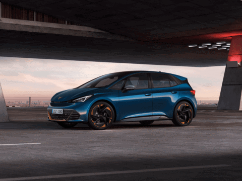 Coche eléctrico azul en estacionamiento urbano bajo puente, con fondo de ciudad y cielo al atardecer.