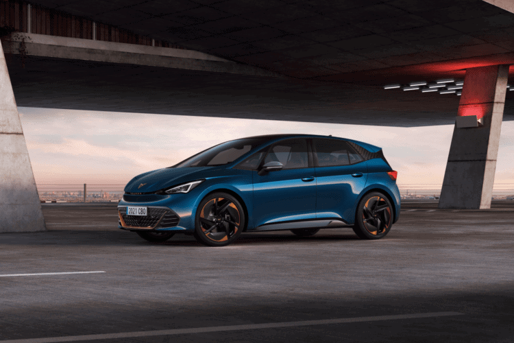 Coche eléctrico azul en estacionamiento urbano bajo puente, con fondo de ciudad y cielo al atardecer.