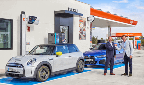 Mini coche eléctrico en estación de carga con dos hombres sonriendo y dándose la mano.