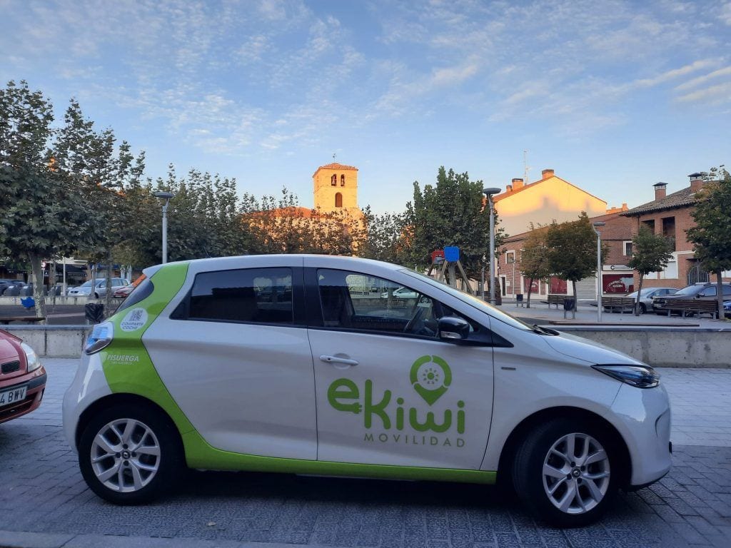Coche eléctrico con logotipo de eKiwí Movilidad en un entorno urbano al atardecer.