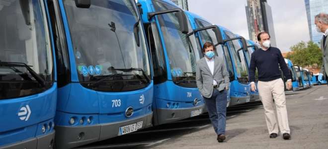 Personas caminando junto a autobuses eléctricos en una parada urbana, usando mascarillas.