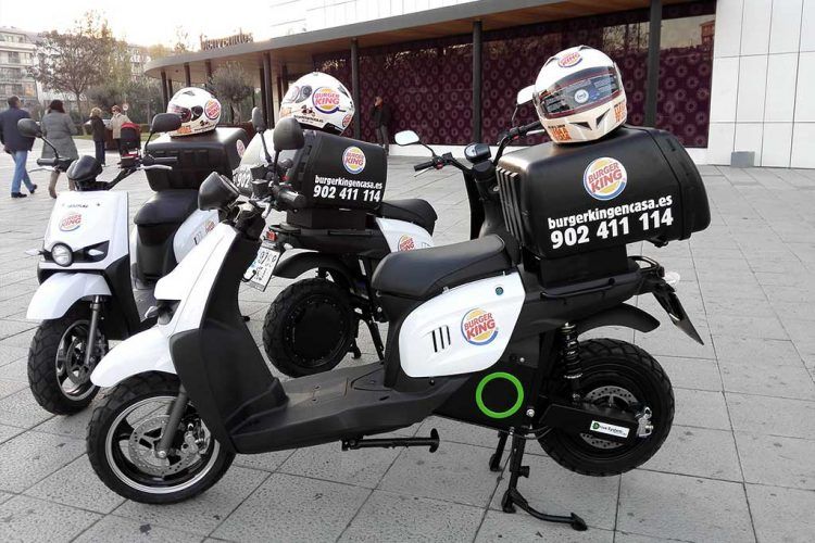Motos eléctricas de reparto con cascos y cajas de Burger King en la ciudad.