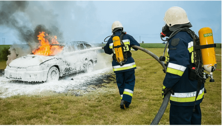 Bomberos apagando un coche en llamas con manguera en campo abierto.